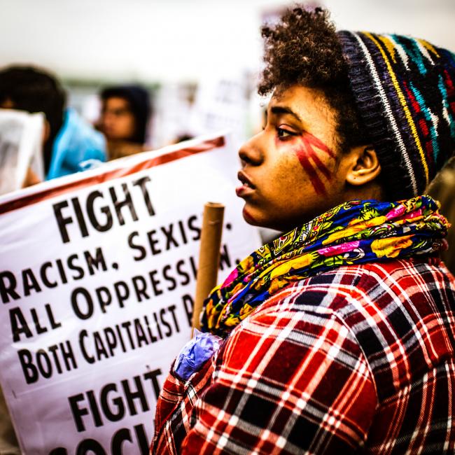 A protester with a sign in the background that reads "Fight Racism. Sexism. All Oppression."